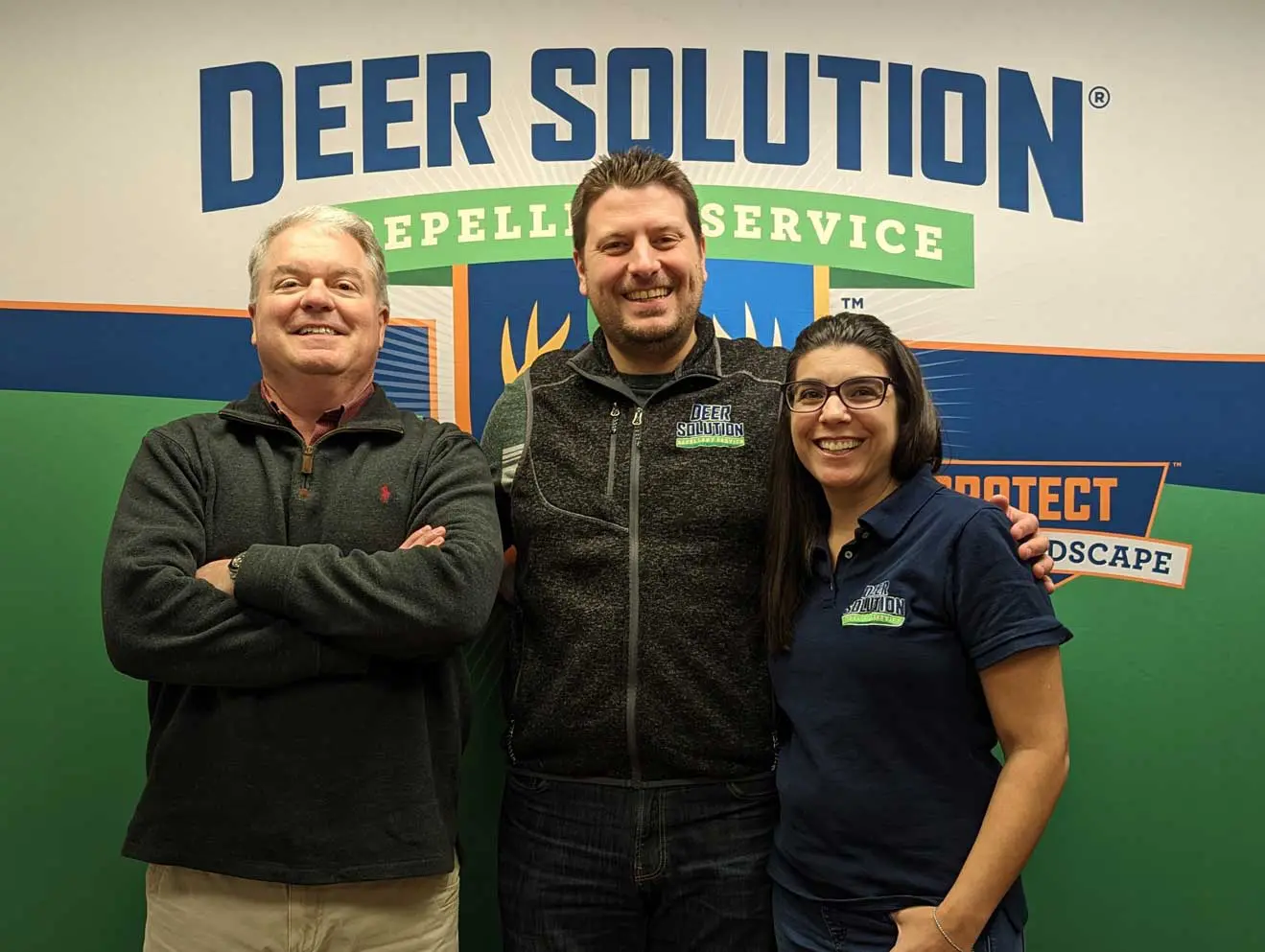 Co-Founders of Deer Solution, Jaime Goodrich on the right and Kris Goodrich in the center, standing with new franchisee Keith Farfone on the left, smiling in front of a Deer Solution logo backdrop.