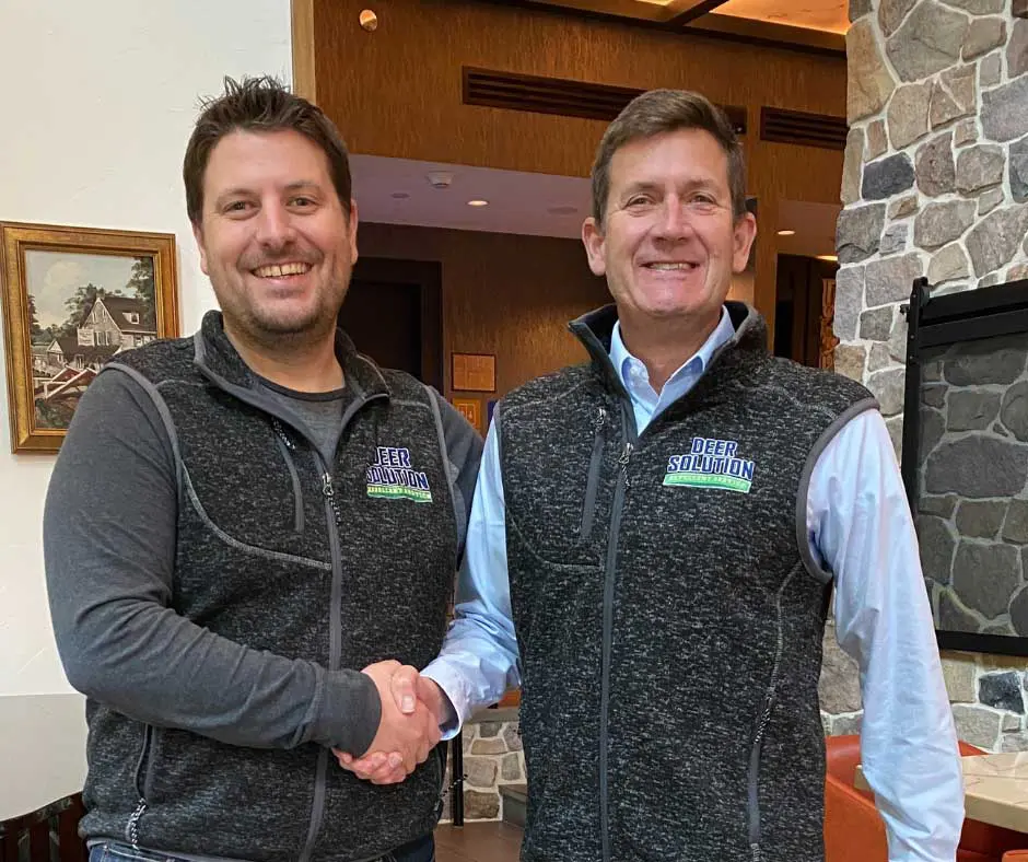 Co-Founder and CEO of Deer Solution, Kris Goodrich, on the left, shaking hands with Joe Garzio, a new franchise owner, both smiling and wearing Deer Solution branded vests in an indoor setting.